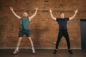 Two men performing jumping jacks in a gym setting, showcasing fitness and energy.